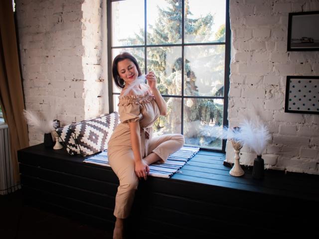 MarieSho with small tits, sitting on black window ledge, wearing beige ruffle top and pants, smiling, touching hair, white brick wall, black window, black and white pillows.