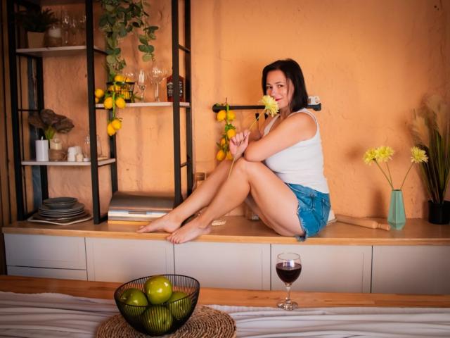 Marie sitzt auf einem Holzboden, hält eine gelbe Blume, trägt ein weißes Tanktop und blaue Shorts, wobei ihr Hintern und ihre Brüste leicht sichtbar sind, neben einem Glas Wein..