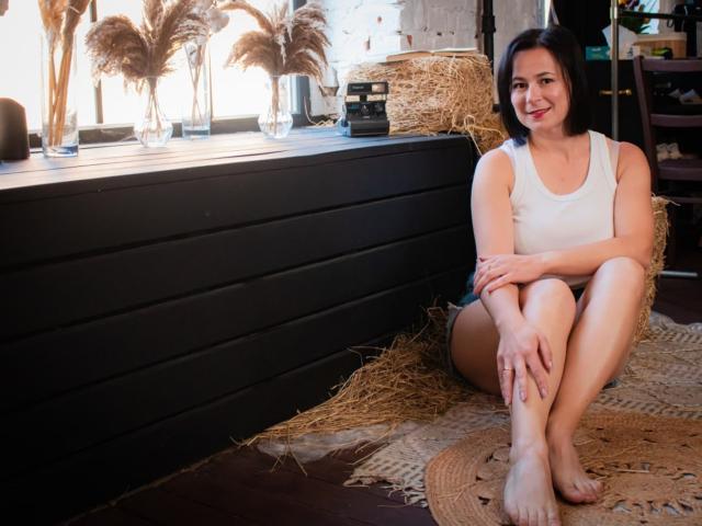 MarieSho with short black hair, white tank top, and no pants, sitting on straw, legs crossed, smiling, barefoot, hands on knees, fluff pampas in glass vases behind.