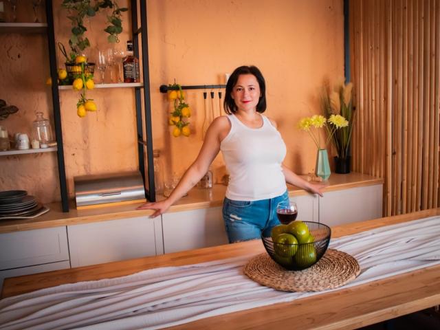 MarieSho with medium tits, wearing a white tank top and ripped blue jeans, stands in a kitchen with hands on the counter, next to a bowl of green apples.