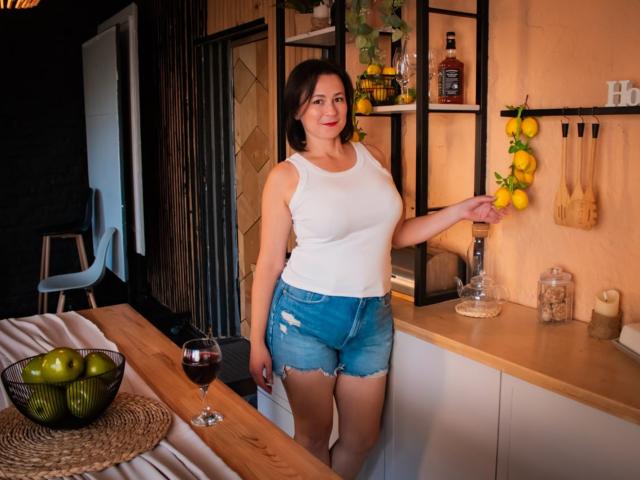 MarieSho with medium tits, white tank top, and blue denim shorts, holding a lemon, standing in a kitchen with a wooden counter, glass of red wine, and green apples.