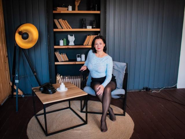 MarieSho mit Brüsten, die von einem hellblauen Pullover bedeckt sind, einem schwarzen Hintern in einem Rock, schwarzen Strumpfhosen, sitzend auf einem Stuhl, an einem Holztisch, mit einer gelben Stehlampe und einem Bücherregal..