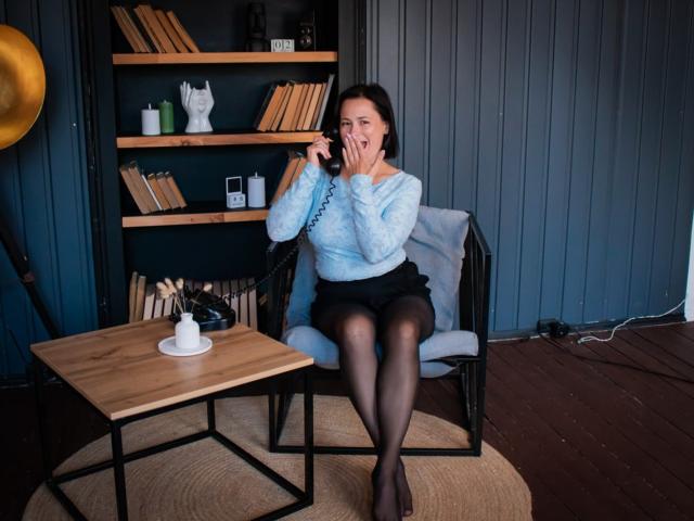 MarieSho with dark hair, blue sweater, black skirt, and black tights, sitting in a chair, talking on a phone, legs crossed, wooden table with a white lamp.