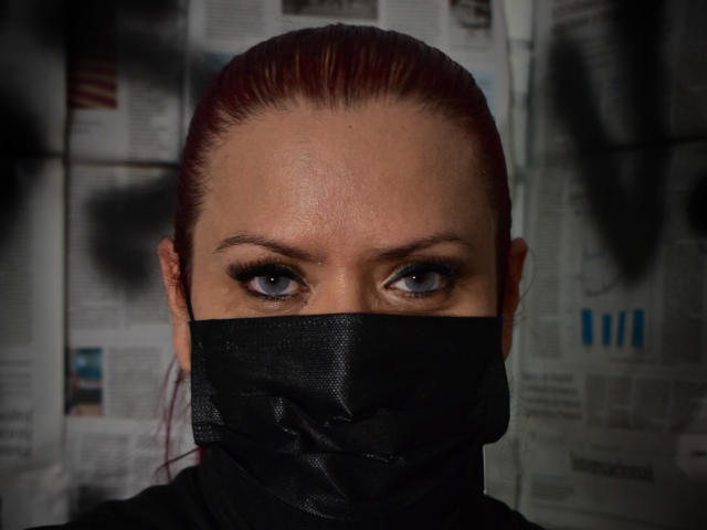 VanesaMoka, com cabelo ruivo, olhos azuis e uma máscara preta cobrindo a parte inferior do rosto, com uma expressão intensa. Suas sobrancelhas estão ligeiramente arqueadas e seu olhar é penetrante..