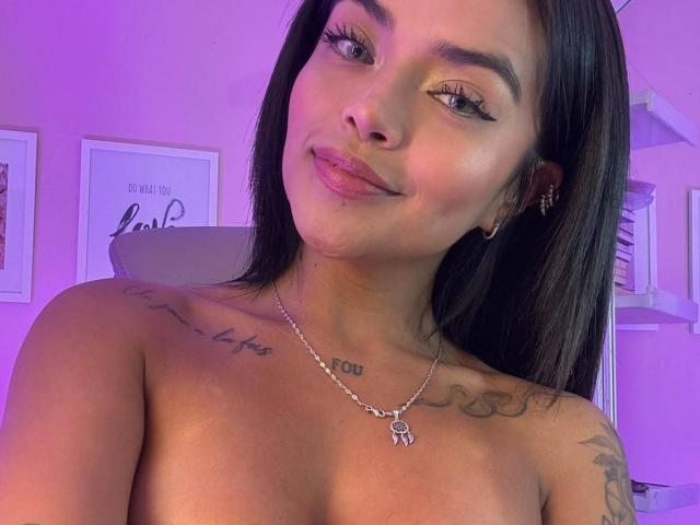 MajoJones tiene una piel marrón media, cabello negro lacio, y está sin ropa, mostrando tatuajes en el pecho y el hombro, con un collar de plata, y una sonrisa sutil..