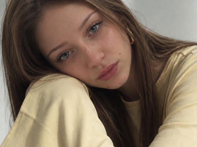 JoelleCalixte has long brown hair, blue eyes, and a small beauty mark on her left cheek. She's wearing a yellow shirt, resting her head on her arm, looking sad.
