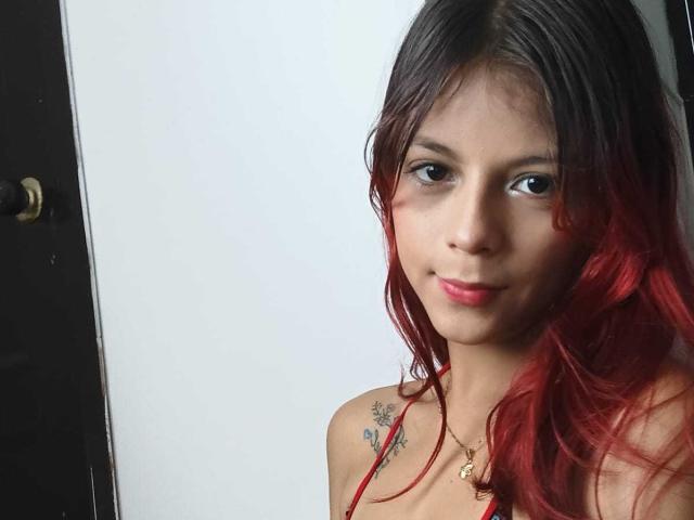 AteneaBlue with red-dyed hair, small tits, tattoos on right shoulder, pink lips, brown eyes, wearing a necklace, standing against a white wall, looking to the left.