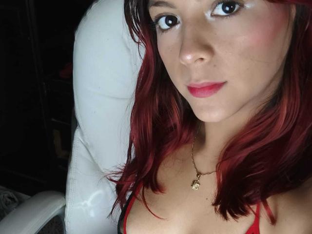 AteneaBlue with red lipstick and red-streaked brown hair, wearing a red top, necklace with a small gold charm, looking into the camera with a slight smile.