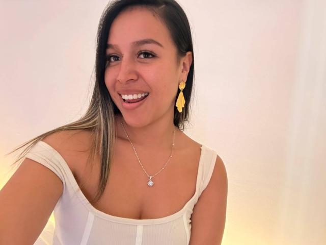 LiaDiane, com seios médios, sorrindo, vestindo uma blusa branca com um ombro descoberto, brincos amarelos e um colar de prata, tem cabelos castanho-claros e um rosto bonito..
