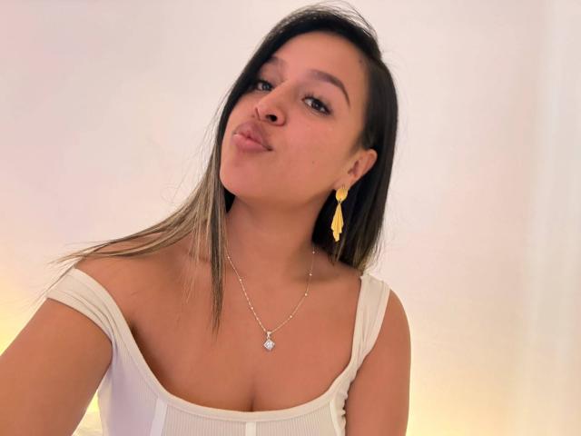 LiaDiane with pouty lips, brown hair, white off-shoulder top, small tits, yellow earrings, and silver necklace, smiling slightly.