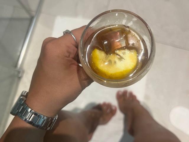 ShilaBrunetti's hand holds a glass with ice, lemon, and whiskey, wearing a silver wristwatch. ShilaBrunetti's bare feet are on a white mat.