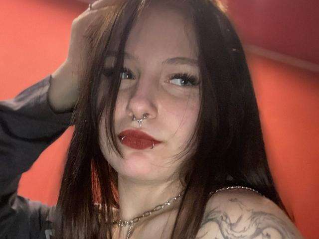 TonetteMasson, com cabelos castanhos longos, olhos azuis, piercings no nariz, piercing no lábio, batom escuro, colar de corrente, braço tatuado, vestindo uma blusa escura, mão no cabelo, fundo vermelho, luz suave..