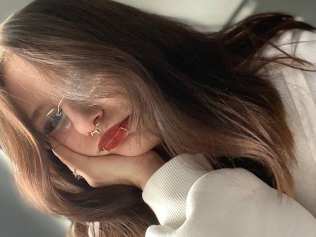 TonetteMasson, con cabello castaño largo, gafas, piercing en la nariz, lápiz labial rojo, suéter blanco, mano en la mejilla, luz suave, cabello ligeramente despeinado, sonrisa sutil, mirando a la cámara, informal..