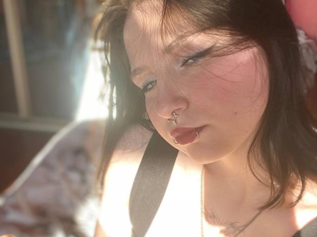 TonetteMasson with sunlit face, brown hair, nose ring, lip ring, and seatbelt, eyes half-closed, wearing floral top, soft light, slight blush, silver necklace, relaxed expression.