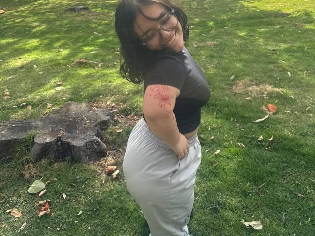 NiniWillow with glasses, black top, gray pants, smiling, showing red tattoo on right arm, curvy butt, standing on grass, tree stump in background, dark wavy hair.