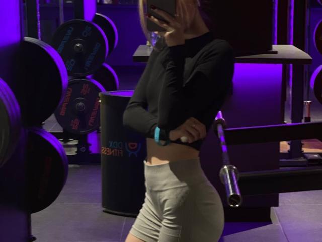 JenetteDubberly in a gym, wearing a black long-sleeve top and tight grey shorts, showing her ass, holding a phone, with purple lighting.