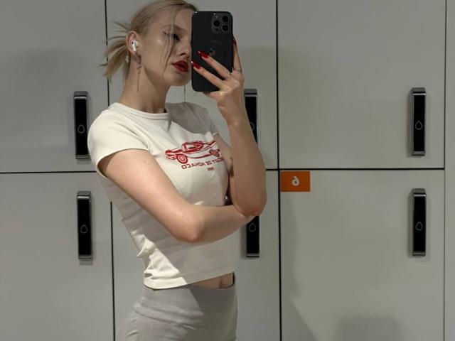 JenetteDubberly with blonde hair, red lipstick, white t-shirt, and grey pants, taking a mirror selfie, showing her tits and holding a black phone.
