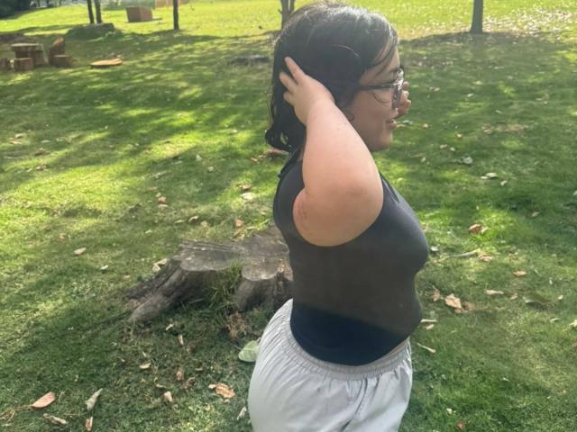 NiniWillow with glasses, dark hair, wearing a black sleeveless top and gray pants, stands in a grassy park, hands in hair, showing her ass and medium tits.