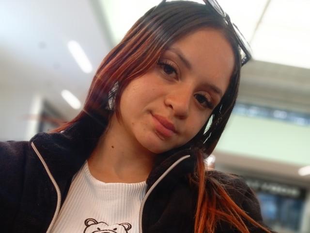 LesleyGray with long brown hair, wearing a black zip-up jacket over a white ribbed shirt, has a small zit on her left cheek, and a slight, confident smirk.
