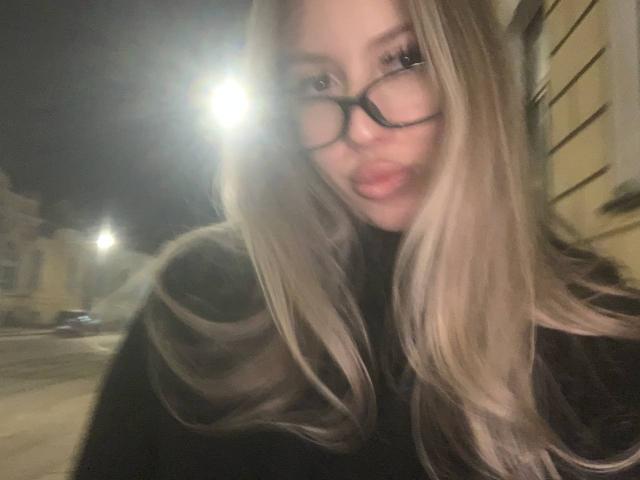 MarcelineConkey with long blonde hair, black glasses, and pouty lips, making a duck face in a blurry nighttime street selfie. Her tits are not visible.