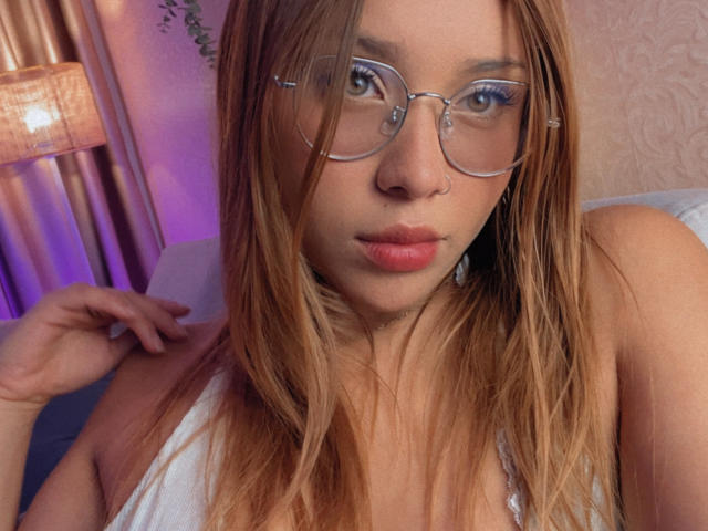 HeraJuno with long blonde hair, wearing clear glasses, and a white top, has pouty pink lips and is looking directly at the camera, showing her small tits.