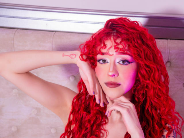 GaiaDoll with bright red curly hair, purple eyeliner, and pink blush, touching her face and neck, showing a small pink tattoo on her forearm. Her tits are partially visible.