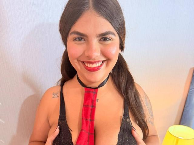 IsaMillerE with pigtails, smiling, wearing a black lace top, red plaid tie, and holding her tits, has tattoos on her shoulders, and red lipstick.