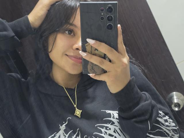 Yurimy mit dunklem Haar, die einen schwarzen Kapuzenpullover trägt, ein Telefon hält, ihre gepflegten Nägel zeigt und eine goldene Halskette mit einem Kreuz trägt, macht ein Selfie im Spiegel..