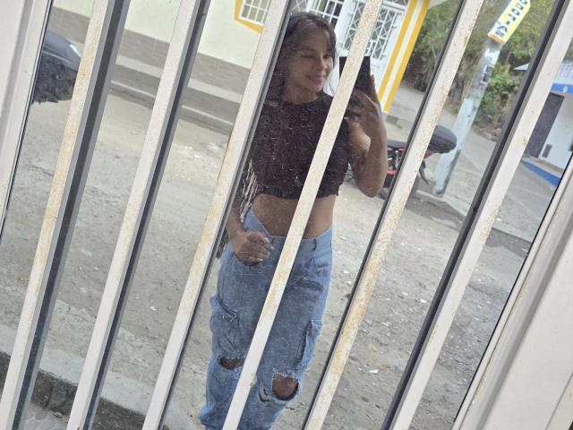 Yurimy, con cabello largo y castaño, vestida con un top corto negro y jeans azules rotos, está de pie frente a una ventana de vidrio sucia, tomando una selfie con el teléfono..