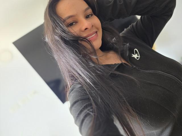 Yurimy met lang zwart haar, lachend, draagt een zwarte hoodie met rits, waarbij haar borsten lichtjes zichtbaar zijn, haar hand in haar haar, bruine ogen, roze lippen, op een selfie..