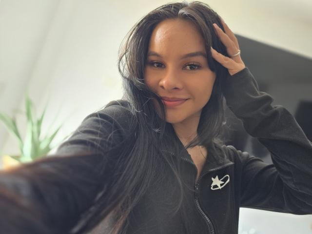 Yurimy, con cabello negro largo, piel morena y una sonrisa encantadora, vestida con una chaqueta negra con cremallera y el logo de Playboy en blanco, sosteniendo su cabello con una mano..