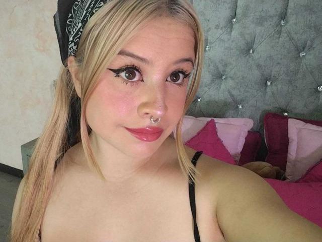 Melani Becker heeft blond haar, een neusring, zwarte eyeliner, roze lippenstift en is topless met zichtbare, kleine borsten. Ze draagt een zwarte haarband in een selfie die in een slaapkamer is gemaakt..