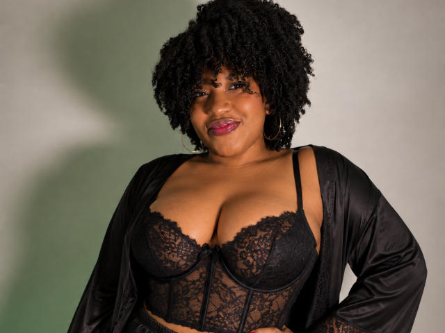 ViaraMoon with curly black hair, dark skin, and pink lipstick, wearing a black lace bra and satin robe, showing her big, round, firm boobs. Smiling confidently.