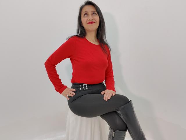 Cyntiia with long black hair, red long-sleeve top, black belt, black skirt, and black knee-high boots, standing with hands on hips, smiling, looking up.