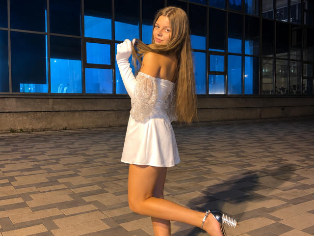 RhonaBells with long brown hair, white off-shoulder dress, and silver high heels, playfully holding her hair, showing her bare ass, standing on a tiled patio at night.