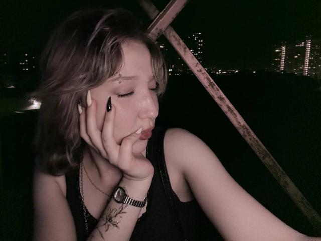 Lorinda Siewert, met kort bruin haar, een zwart mouwloos topje, zwarte nagels en een tatoeage op haar pols, met een frons en haar hand op haar wang, met stadslichten op de achtergrond, onder een roestige metalen constructie..