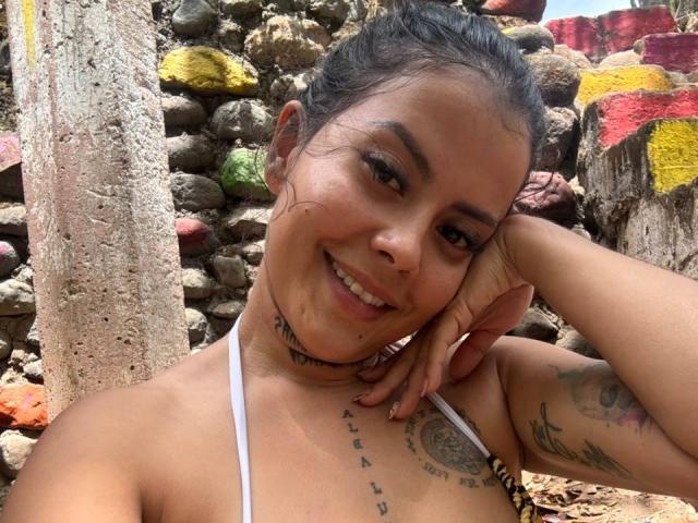 ElectraRoss with tan skin, dark hair in a messy bun, smiling, white halter top, tattoos on neck and chest, arm tattoo, hand on cheek, stone wall with colorful paint.