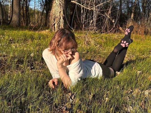 AnnadelEmeralda, lying on grass, wearing a white shirt, black leggings with pink paw prints, and pink-tinted hair, with her ass up and legs in the air.