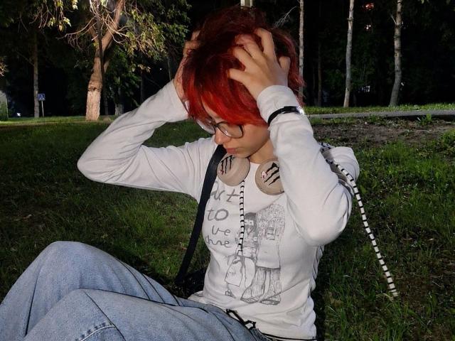 AnnadelEmeralda with red hair, glasses, white long-sleeve shirt, and blue jeans, sitting on grass, hands in hair, looking down, headphones around neck, dark forest background.