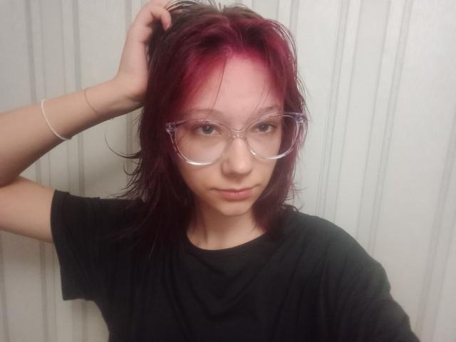 AnnadelEmeralda with pink-dyed hair, clear glasses, black shirt, and wrist bracelet, holding hair, looking at camera. Her face is smooth, with a slightly serious expression.