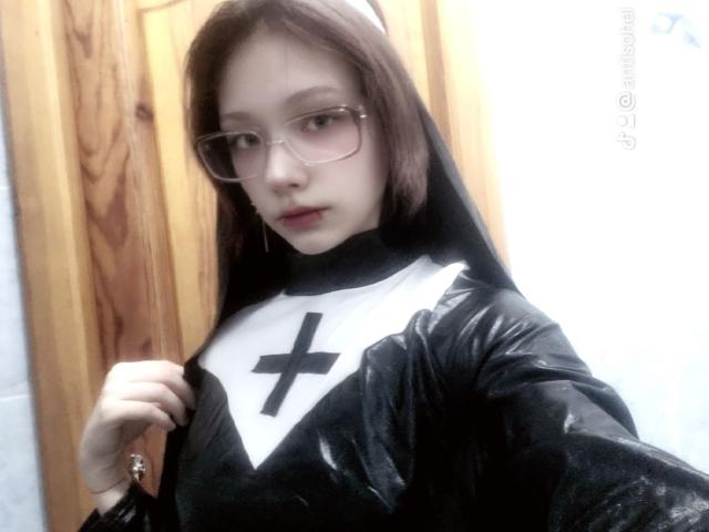AnnadelEmeralda in a black and white nun outfit, holding it open to show a large black cross on her chest, with glasses, brown hair, and a wooden door.
