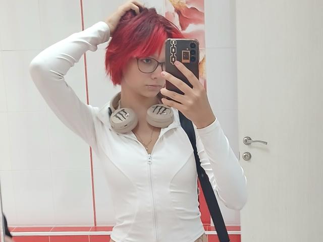 AnnadelEmeralda with red hair, glasses, white zip-up, headphones, mirror selfie, hand in hair, holding phone, white tiles, door, phone case with chain design.