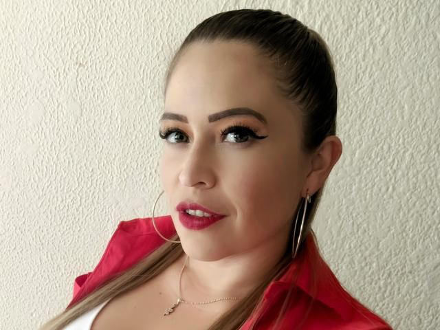 ImEmilyHanks has red lipstick, hoop earrings, and a red shirt, with brown hair pulled back, wearing eyeliner, and a necklace, looking at the camera with a slight smile.