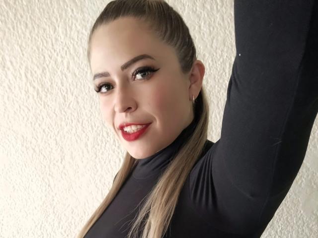 ImEmilyHanks with a sleek ponytail, wearing a black long-sleeve top, smiling with red lipstick, arm raised, showing off her tits subtly, against a textured white wall.