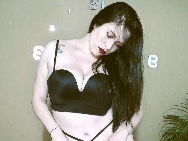 ImEmilyHanks with pale skin, long black hair, dark lipstick, black bra, and black panties, looking down, showing her big tits, and a small tattoo on her left shoulder.