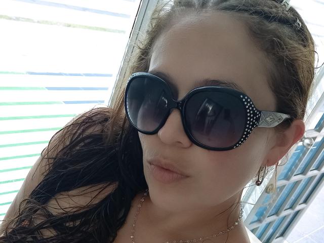 ImEmilyHanks with wet brown hair, wearing black polka-dotted sunglasses, gold earrings, and a necklace, has pouty lips and fair skin. Her boobs are partially visible.