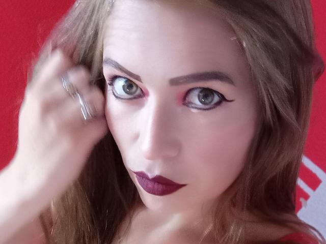 ImEmilyHanks has pale skin, green eyes with bold eyeliner, and dark purple lipstick. She's touching her hair with a silver ring, looking intense. Her tits are not visible.