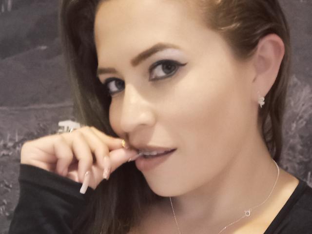 ImEmilyHanks with manicured nails, black eyeliner, and subtle smile, touching her lip, wearing a black top and silver necklace, showing her tits slightly in the frame.