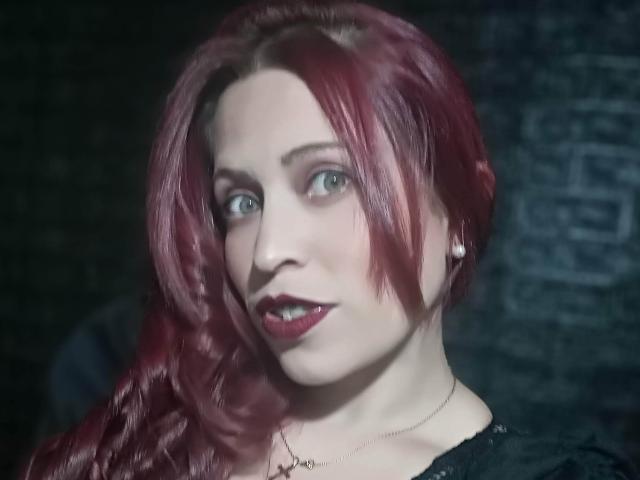 ImEmilyHanks has red hair, pale skin, green eyes, and red lipstick. She's wearing a black top with a necklace. Her ass is not visible. Her tits are small.