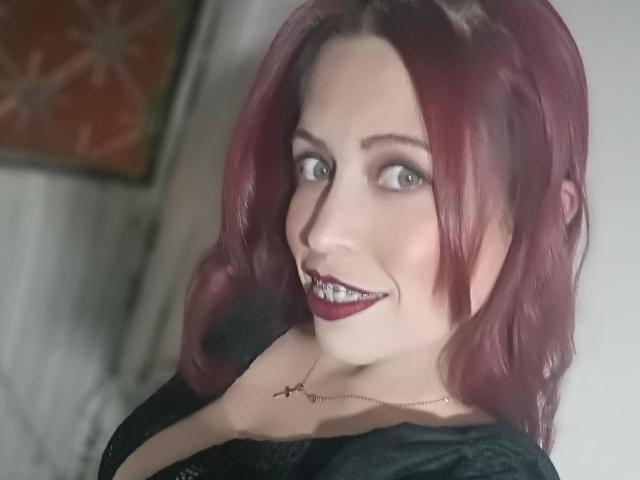 ImEmilyHanks with red hair, blue eyes, and dark lipstick, smiling with her hand near her tits, wearing a black top and gold necklace.